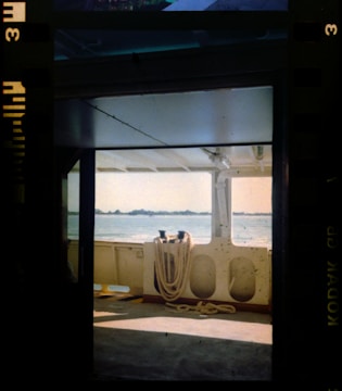 A view from inside a boat looking out towards a calm body of water. The interior features ropes coiled neatly on hooks along the wall and the scene is framed by sections of the boat's structure. Natural light filters through, casting shadows and highlighting the industrial design.