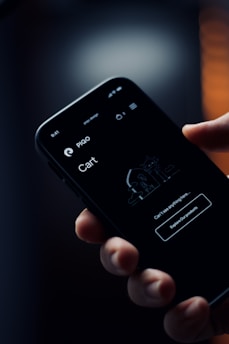 A hand is holding a smartphone with a dark-themed screen displaying a shopping cart interface. The screen shows text reading 'Can't see anything here...' and a button labeled 'Explore our products'. The background appears blurry with warm, soft lighting.