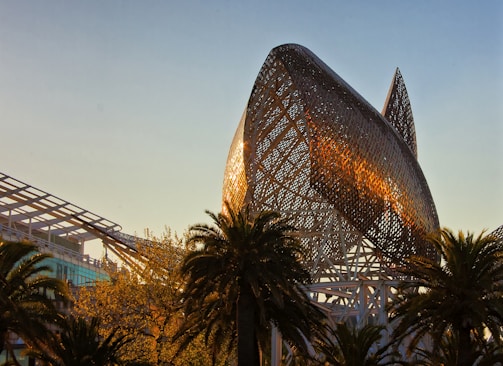A large, metallic, abstract sculpture resembles the shape of a fish, with a lattice-like structure that catches the warm glow of the setting sun. Palm trees and modern architectural elements surround the sculpture, creating a juxtaposition of nature and contemporary design.