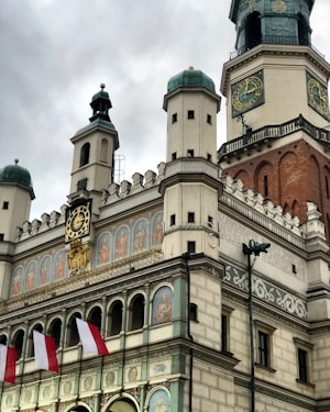 A historic building with ornate architectural features, including multiple towers with teal domes. Detailed frescoes line the fa&ccedil;ade beneath large clocks, showcasing intricate artwork. Several red and white flags are displayed in the foreground, adding a splash of color against the predominantly muted stone walls.
