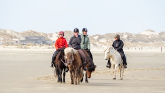 Four people wearing helmets are riding horses along a sandy beach, with dunes visible in the background. The riders are dressed in casual riding attire and appear to be enjoying a leisurely ride. The setting is calm and expansive.
