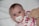 A baby with bright eyes is lying on a white surface, using a pacifier with a small duck design. The baby is wrapped in a blanket with colorful floral patterns in pink, brown, and orange. A soft teddy bear is positioned beside the baby.