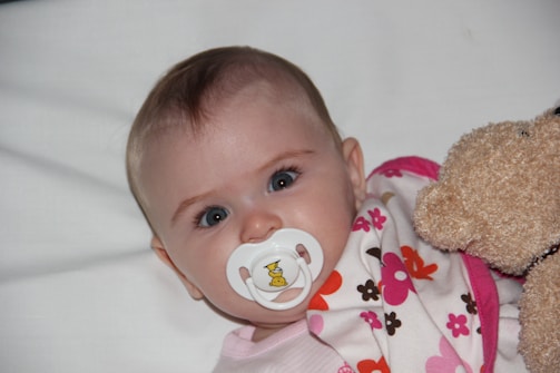 A baby with bright eyes is lying on a white surface, using a pacifier with a small duck design. The baby is wrapped in a blanket with colorful floral patterns in pink, brown, and orange. A soft teddy bear is positioned beside the baby.