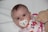 A baby with bright eyes is lying on a white surface, using a pacifier with a small duck design. The baby is wrapped in a blanket with colorful floral patterns in pink, brown, and orange. A soft teddy bear is positioned beside the baby.
