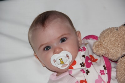 A baby with bright eyes is lying on a white surface, using a pacifier with a small duck design. The baby is wrapped in a blanket with colorful floral patterns in pink, brown, and orange. A soft teddy bear is positioned beside the baby.