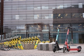 A row of yellow rental bicycles is lined up neatly on a paved area in front of a modern building with large glass windows. Next to them are two electric scooters, one teal and one red, positioned upright. Large stone blocks are scattered nearby, and the building windows reflect parts of the surrounding urban environment.