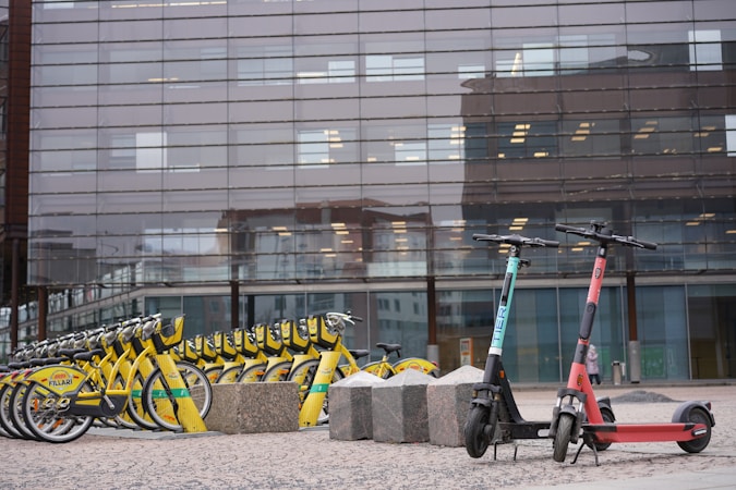 A row of yellow rental bicycles is lined up neatly on a paved area in front of a modern building with large glass windows. Next to them are two electric scooters, one teal and one red, positioned upright. Large stone blocks are scattered nearby, and the building windows reflect parts of the surrounding urban environment.