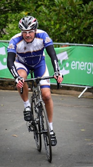 A cyclist in racing attire rides a road bike on a paved path. He wears a helmet and glasses, suggesting participation in a competitive event. Green banners with text are visible in the background, indicating sponsorship or event signage. The setting includes lush greenery.