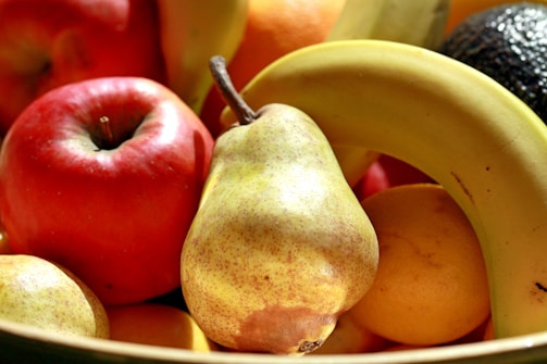 A close-up of a vibrant assortment of fruits, including a red apple, a ripe banana, a pear, and citrus fruits. The arrangement highlights the natural textures and colors under bright lighting, creating a vivid and appetizing display.