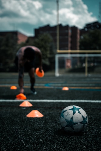 A person is engaged in a soccer training session on an outdoor field. The individual is focused on maneuvering through orange cones arranged on the grass. In the foreground, a soccer ball rests on the ground. There are buildings and trees in the background under a partly cloudy sky.
