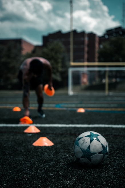 A person is engaged in a soccer training session on an outdoor field. The individual is focused on maneuvering through orange cones arranged on the grass. In the foreground, a soccer ball rests on the ground. There are buildings and trees in the background under a partly cloudy sky.