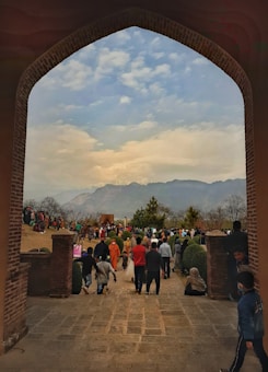 A large crowd of people is gathered in a garden setting, framed by an arched brick entrance. The background features a mountain range under a partly cloudy sky. People are walking, standing, and wearing varied clothing, suggesting a casual, social atmosphere.