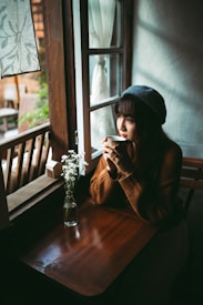 A person is sitting by a window holding a cup, wearing a beret and sweater. A vase with small white flowers is on the wooden table beside them. The room has a calm and cozy atmosphere with natural light streaming in.
