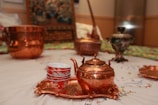 A copper teapot and a stack of red and white patterned teacups are placed on a copper tray. These are set on an intricately designed fabric. In the background, additional copper and decorative pots can be seen, alongside other ornamental objects.