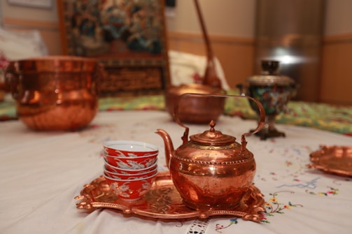 A copper teapot and a stack of red and white patterned teacups are placed on a copper tray. These are set on an intricately designed fabric. In the background, additional copper and decorative pots can be seen, alongside other ornamental objects.