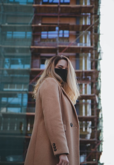 A person in a brown coat stands in front of a building under construction, with scaffolding visible. They are wearing a black mask and have long, blonde hair. The background shows glass windows reflecting light.