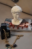 On a light-colored surface, there is a black perfume bottle adorned with a gold chain. Next to the bottle, there is a magazine with a cover featuring a person's face and the title 'Marie Claire'. A white bust sculpture candle is placed on top of the magazine.