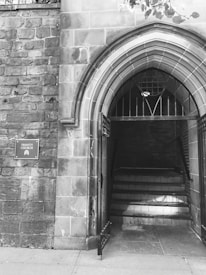 An entrance to a building with Gothic architectural features, showcasing a pointed archway and an open gate. A sign beside the entrance reads 'Trinity Church', indicating the location.