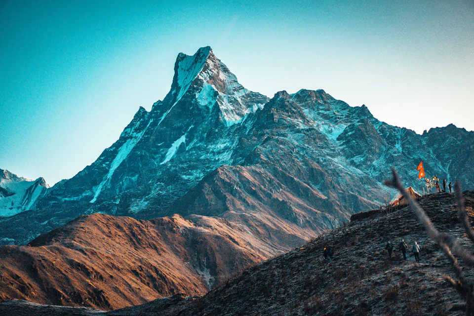 The image depicts a majestic mountain range with snow-capped peaks and a clear blue sky. The mountains, painted in shades of brown and gray, rise high into the sky, creating a sense of grandeur and scale. The perspective is from a low angle, looking up at the towering mountains, which enhances their imposing presence. In the foreground, there are several people scattered across the landscape, adding a touch of life to this otherwise serene scene.
