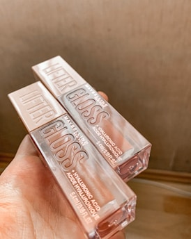 Two tubes of lip gloss are held in a hand, featuring clear packaging with visible branding text. The gloss is labeled as 'Lifter Gloss' and contains hyaluronic acid. The background is blurred, highlighting the products.
