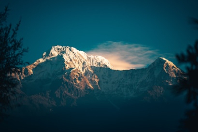 Snow-capped mountains are illuminated by soft, warm sunlight, contrasting with the deep blue sky. Wispy clouds hover around the peaks, adding a sense of serene majesty. Dark silhouettes of foliage frame the scene, enhancing the grandeur of the landscape.