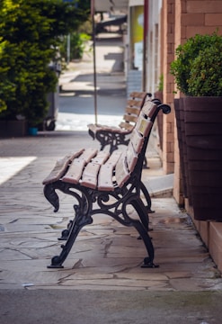 Wooden benches with ornate metal legs line a stone-paved walkway. A large planter with lush green shrubs is visible nearby, and the background showcases a sunlit outdoor area with shaded spots.