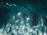 Waves crashing in a swirling pattern with varying shades of blue and white foam visible on the ocean surface, conveying motion and energy.