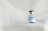 A small glass vial with a blue cap and label is positioned on a soft, light-colored surface. The label indicates it is a SARS-CoV-2 Protein Subunit Vaccine for COVID-19.