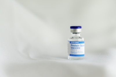 A small glass vial with a blue cap and label is positioned on a soft, light-colored surface. The label indicates it is a SARS-CoV-2 Protein Subunit Vaccine for COVID-19.