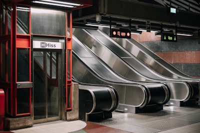 A subway station features an elevator with a 'Hiss' sign next to three parallel escalators. The escalators are metallic and clean, with overhead signs indicating directions. The area is well-lit with a red and metallic color scheme, and the environment appears modern and minimalistic.