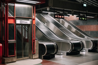 A subway station features an elevator with a 'Hiss' sign next to three parallel escalators. The escalators are metallic and clean, with overhead signs indicating directions. The area is well-lit with a red and metallic color scheme, and the environment appears modern and minimalistic.