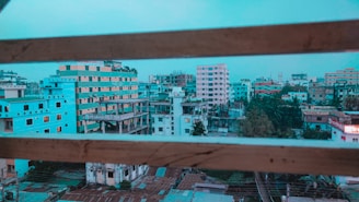 A cityscape featuring multiple buildings of various heights and colors, viewed through a set of horizontal window bars. The buildings range from unfinished structures to completed apartment complexes, with a mix of architectural styles. Some are painted in bright colors, such as turquoise and peach. The scene also includes greenery from trees scattered among the buildings, under a sky that appears to be slightly overcast.
