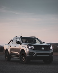 A silver pickup truck is parked on a rough terrain with its headlights on. The vehicle is positioned at an angle, displaying its robust design and dark accents. The background shows a dimly lit sky, creating a contrast against the reflective surface of the truck.