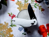 A folding knife with a white handle and a black blade is placed on top of a glass of milk. The background features a white tablecloth with a floral design consisting of colorful flowers and leaves.
