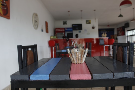 A cozy cafe with a focus on a wooden table painted in black, red, and blue. The table is set with a decorative centerpiece made of sticks. In the background, there is a red counter with high chairs and some wall decorations. The lighting is soft, coming from ceiling lamps.
