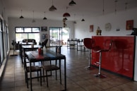 A dining area with several tables and chairs arranged neatly. The room features a tiled floor and large windows that let in natural light. There is a striking red counter with two modern bar stools. Decorative items, including wall clocks and art, adorn the white walls. Pendant lights hang from the ceiling.