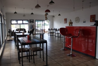 A dining area with several tables and chairs arranged neatly. The room features a tiled floor and large windows that let in natural light. There is a striking red counter with two modern bar stools. Decorative items, including wall clocks and art, adorn the white walls. Pendant lights hang from the ceiling.