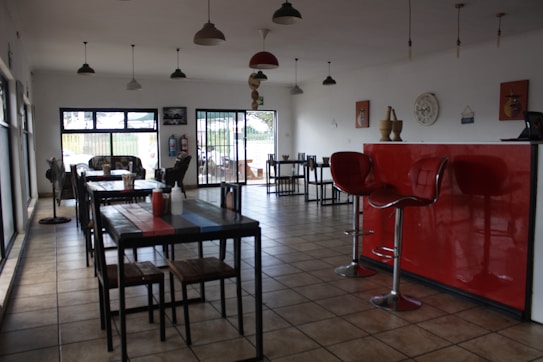 A dining area with several tables and chairs arranged neatly. The room features a tiled floor and large windows that let in natural light. There is a striking red counter with two modern bar stools. Decorative items, including wall clocks and art, adorn the white walls. Pendant lights hang from the ceiling.