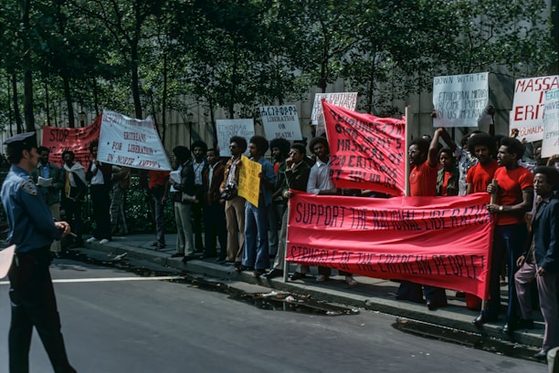 A group of people is gathered on a street for a protest, holding various signs and banners. The banners contain messages supporting the Eritrean liberation struggle. The protesters are standing together near a line of trees, and one police officer is observing them from the street.