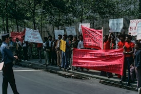 A group of people is gathered on a street for a protest, holding various signs and banners. The banners contain messages supporting the Eritrean liberation struggle. The protesters are standing together near a line of trees, and one police officer is observing them from the street.