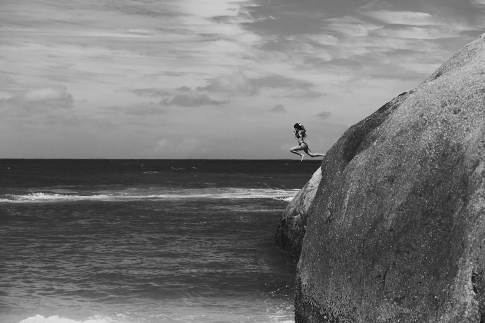 grayscale photo of man sitting on rock near sea