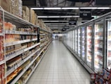 An aisle in a supermarket with shelves on the left displaying a variety of packaged goods, including sauces and snacks. On the right, there is a long row of freezer units containing frozen products. The floor is tiled, and there is bright artificial lighting from overhead.