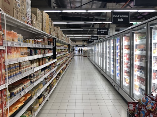 An aisle in a supermarket with shelves on the left displaying a variety of packaged goods, including sauces and snacks. On the right, there is a long row of freezer units containing frozen products. The floor is tiled, and there is bright artificial lighting from overhead.