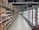 An aisle in a supermarket with shelves on the left displaying a variety of packaged goods, including sauces and snacks. On the right, there is a long row of freezer units containing frozen products. The floor is tiled, and there is bright artificial lighting from overhead.