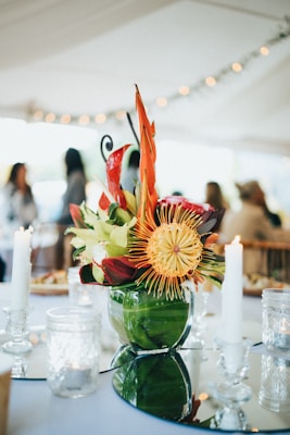 A beautifully arranged centerpiece combining vibrant tropical flowers in red, orange, and yellow within a green glass vase. The arrangement is placed on a reflective surface with softly lit candles surrounding it, creating a warm and elegant ambiance. In the background, blurred figures of people are visible, suggesting a social gathering or event under a white canopy adorned with string lights.