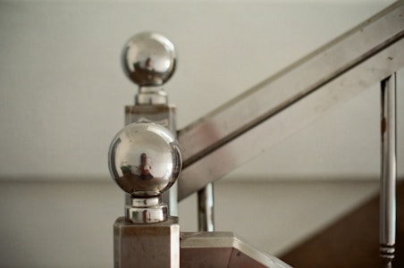 A close-up view of a metallic staircase with round reflective finials. The metal surface is highly polished, showing reflections from the surroundings. The image focuses on the design details of the staircase, highlighting the clean lines and shiny texture.