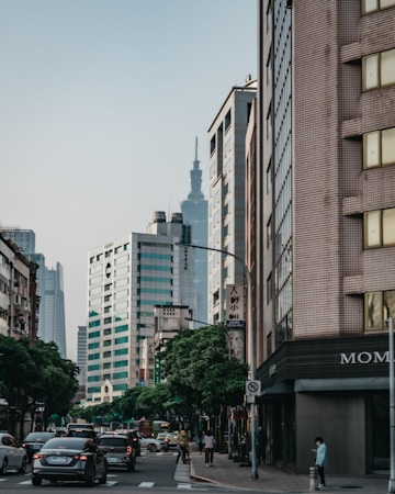 An urban scene featuring a street lined with modern high-rise buildings. In the distance, a tall tower is visible amidst the slightly overcast sky. Cars are moving through the intersection, and trees line the sidewalk. Several pedestrians are walking along the street, and there is a sense of everyday city life.