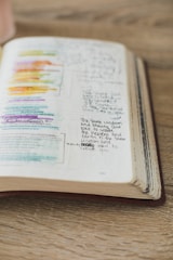 An open book lies on a wooden surface with handwritten notes and highlighted text in various colors. The pages display a mix of printed text and personal annotations made by someone studying or reflecting on the content.