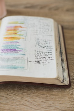 An open book lies on a wooden surface with handwritten notes and highlighted text in various colors. The pages display a mix of printed text and personal annotations made by someone studying or reflecting on the content.