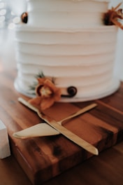 A close-up of a tiered white cake with simple, elegant decorations. The cake has a textured finish and is adorned with small floral arrangements featuring orange flowers and other natural elements. It is situated on a wooden board with a gold cake knife and server beside it.